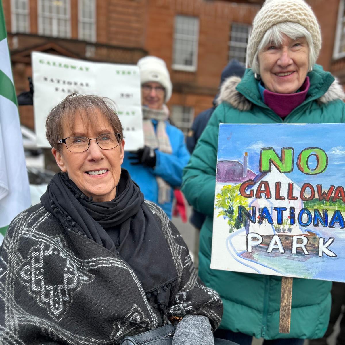 Two women at a protest. One has grey hair and is wearing a cream-coloured woolly hat and a green quilted jacket with a fluffy hood. She has a maroon polo-neck underneath. She is holding a banner that has a painting of a landscape on it with the words No Galloway National Park. The other woman,  Liz Hitschmann, has short, darker hair and glasses. She is wearing a black scarf and a patterned grey shawl over a grey coat.