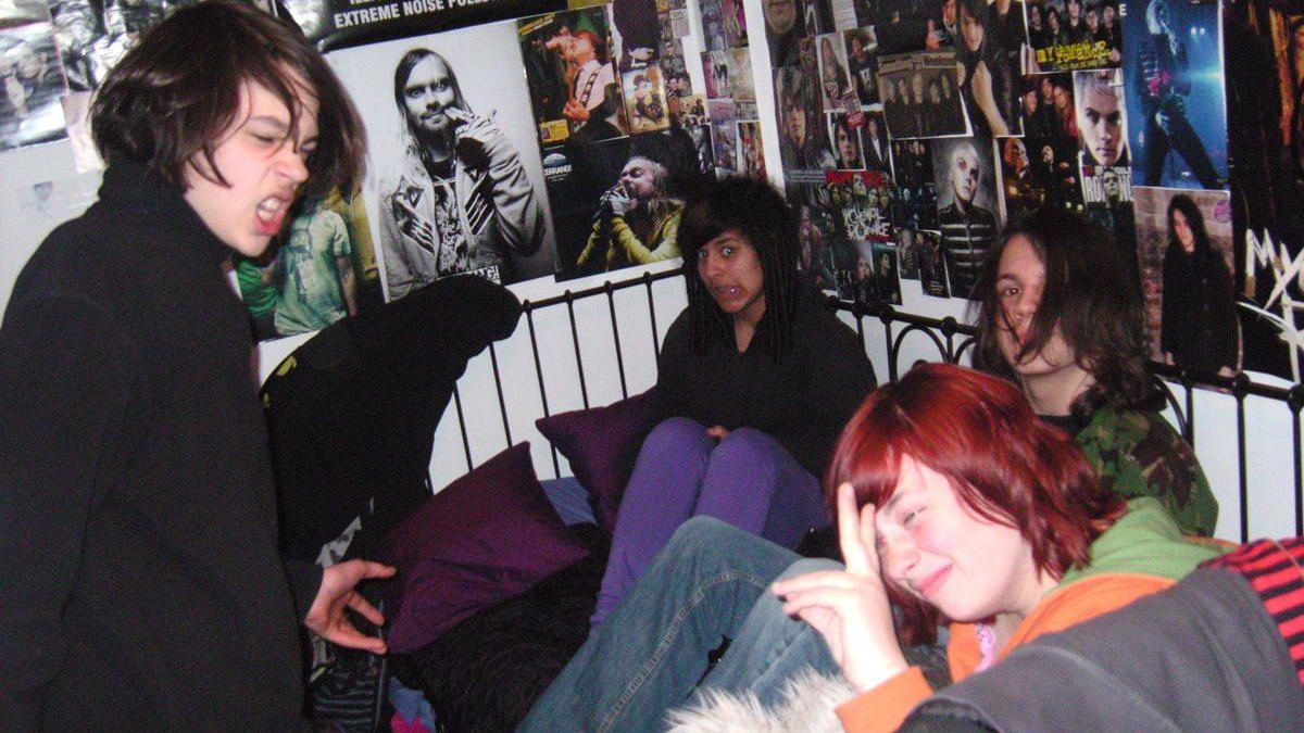 Four people on a black metal frame bed surrounded by posters of emo bands on the wall. A boy on the left has long brown hair and a black jacket, in the middle is a girl with black hair and a fringe. She has a black hoodie and purple trousers. To the right of her is a boy with long hair wearing a camouflage top and jeans. In the front right is a person with red hair motioning a peace sign with her fingers. 