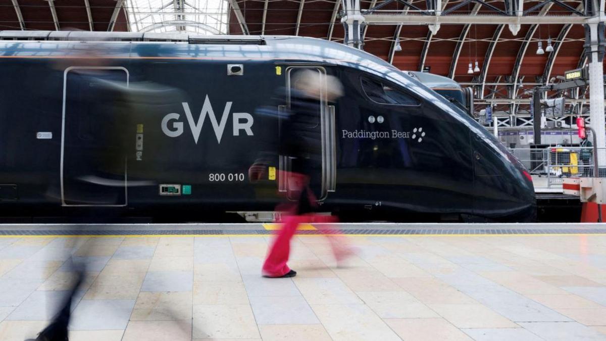 A dark green Great Western Railway train sitting in London Paddington. The train is called Paddington Bear and there is a pedestrian wearing red trousers walking in front of the train. 