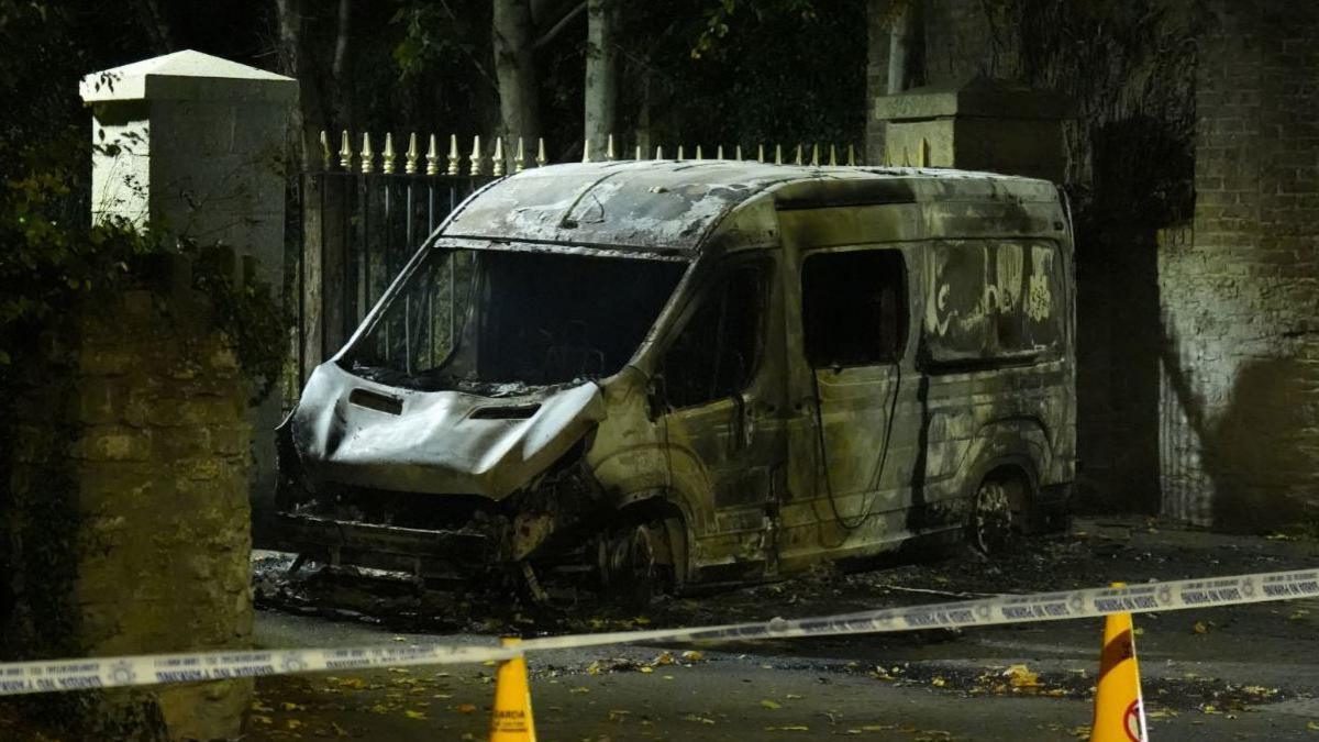 The wreckage of a burned out garda van in Dublin. It is windowless and has scorch marks. There is police tape nearby and yellow cones.