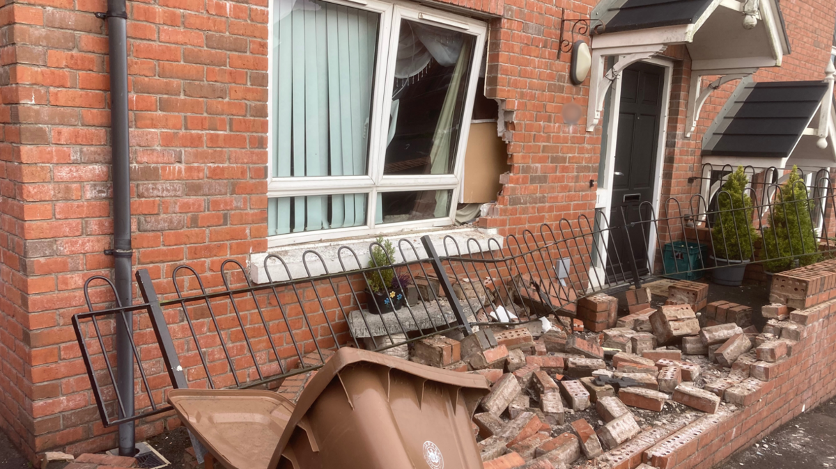 A red brick house with a damaged red brick wall and a damaged black gate in front of it. There is a brown bin fallen over to the front of the wall. 