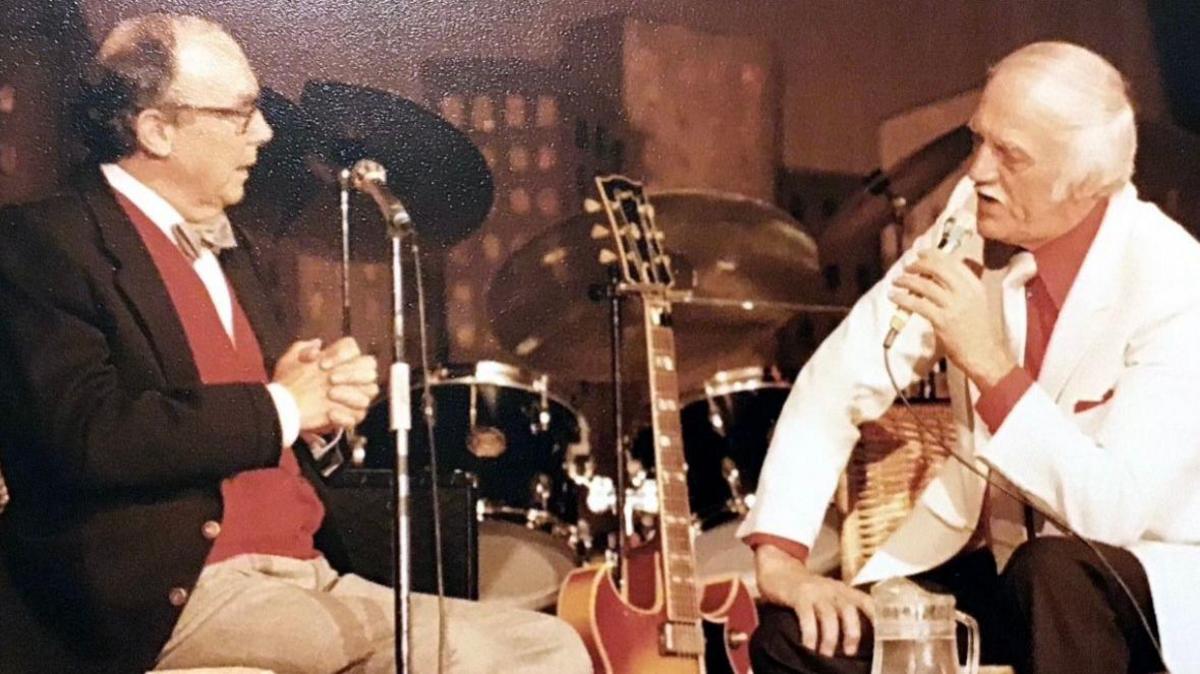 Eric Morecambe sits on a stage, surrounded by music instruments such as a guitar and a drum kit, as he turns to speak into a microphone on a stand to an older man, who is speaking into a microphone. Morecambe is wearing dark glasses, beige bow tie, red cardigan, brown blazer and cream trousers, and the other man is wearing a white jacket, a red shirt, white tie and dark brown trousers.