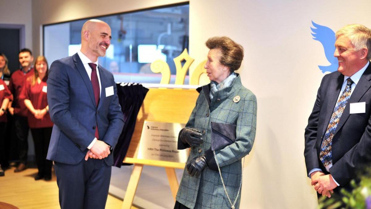 Princess Anne speaking to two men in suits speaking in front of silver board with black writing on. 