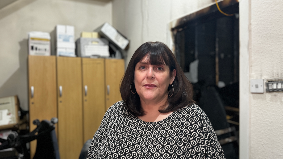 A woman in a white and black patterned shirt stands in front of a burnt-out door leading to a damaged office.