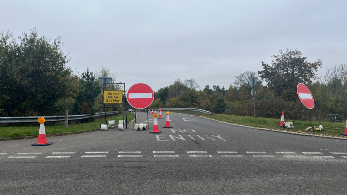 One lane of a slip road has been cordoned off by two large no entry signs, red circles with a horizontal white line in the centre. There are also cones cordoning off the slip road in a line from the font of the image around the slip road, until they are out of sight. A yellow sign with black writing reading 'Do not follow sat nav' has also been put on the blocked slip road. On the road, letters written in white paint read 'No Entry'. 