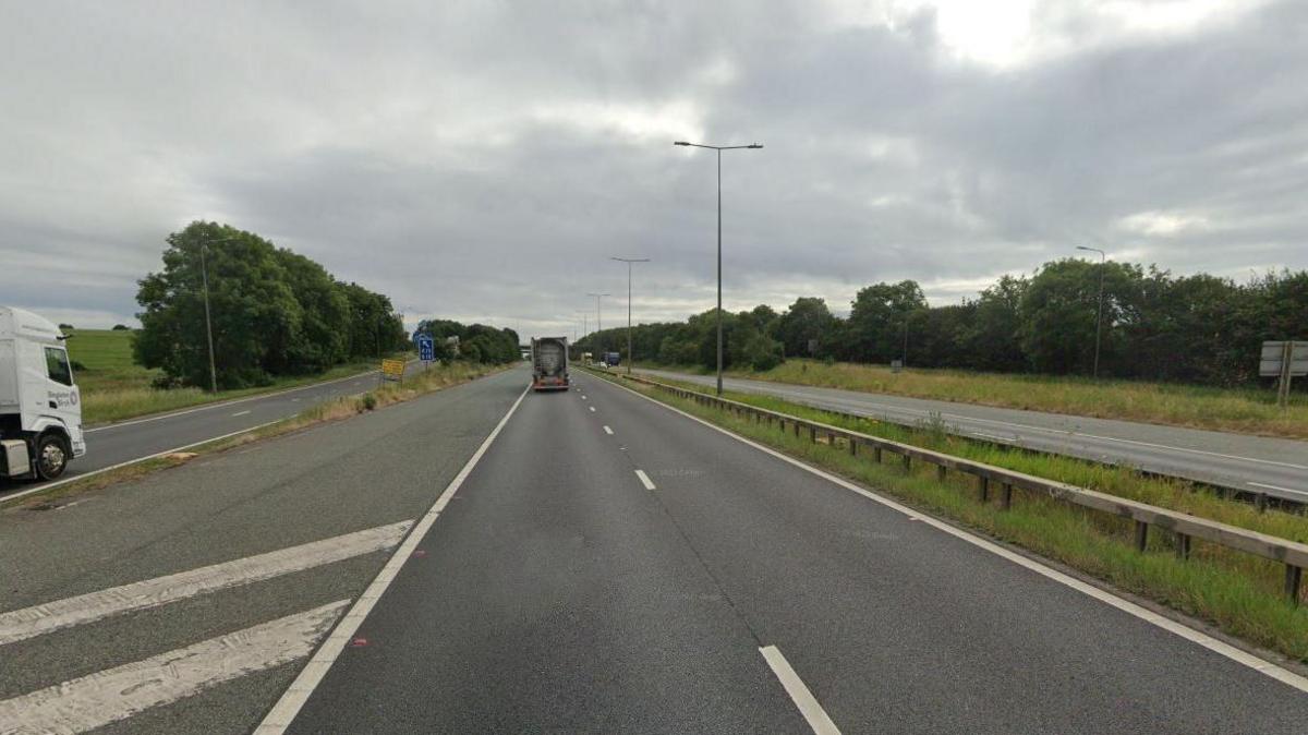 Google road view of the M180 near Barnetby. Trees are seen either side of the road with two lorries seen on the road.