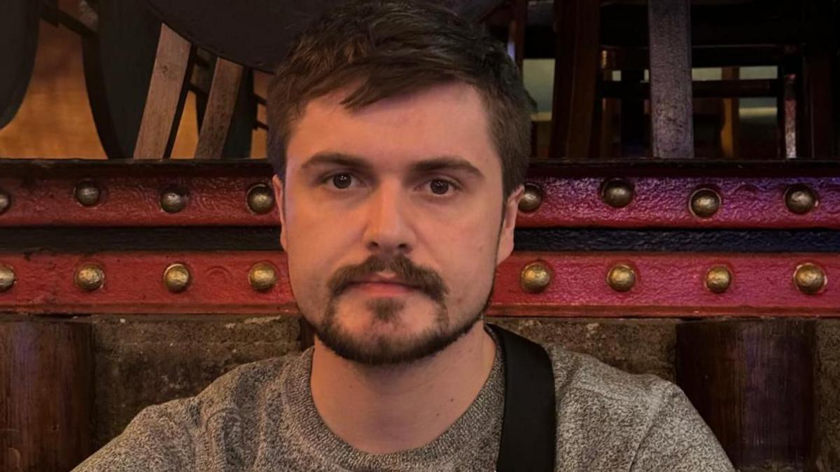 Osian looking at the camera. He is wearing a grey sweater with a black bag strap across his chest. Behind him is a wooden panel with a red horizontal bar running across the top of it with big gold bolts along it. 