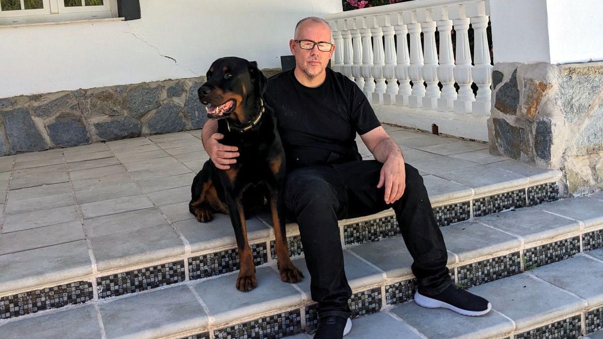 Ryan O'Meara sitting on a step outside a house with his dog. He wears a dark t-shirt and jeans and black trainers with white soles. He is wearing glasses. His dog is large, brown and black and with big teeth. 