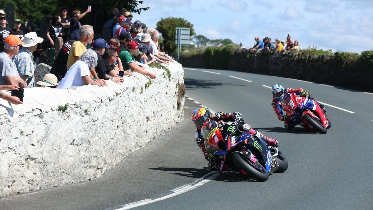 Davey Todd ahead of Dean Harrison on the road as the round a bend while spectators watch the action from behind a white painted stone wall.