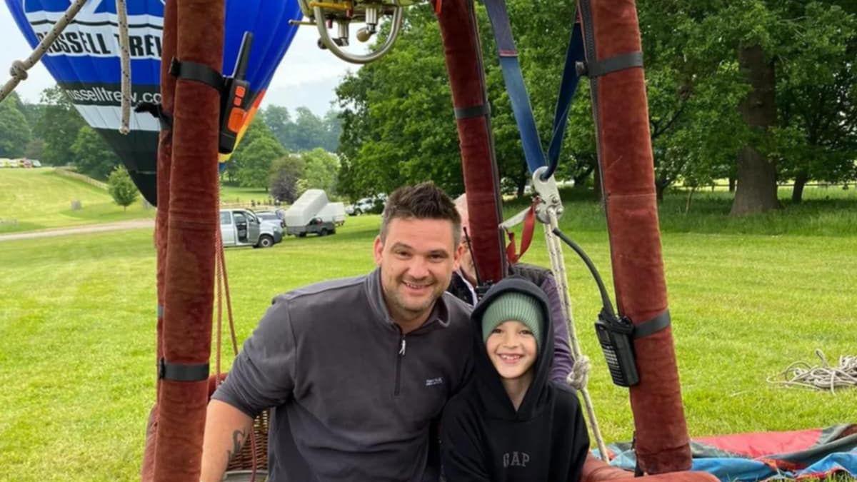 Oliver Surridge, in a hot air balloon, with a child. Oliver has on a grey top, the child has a green hat on, a black hoodie and they are both looking at the camera and smiling. The hot air balloon has red columns and there is a balloon behind him. Grass and trees are behind him also.