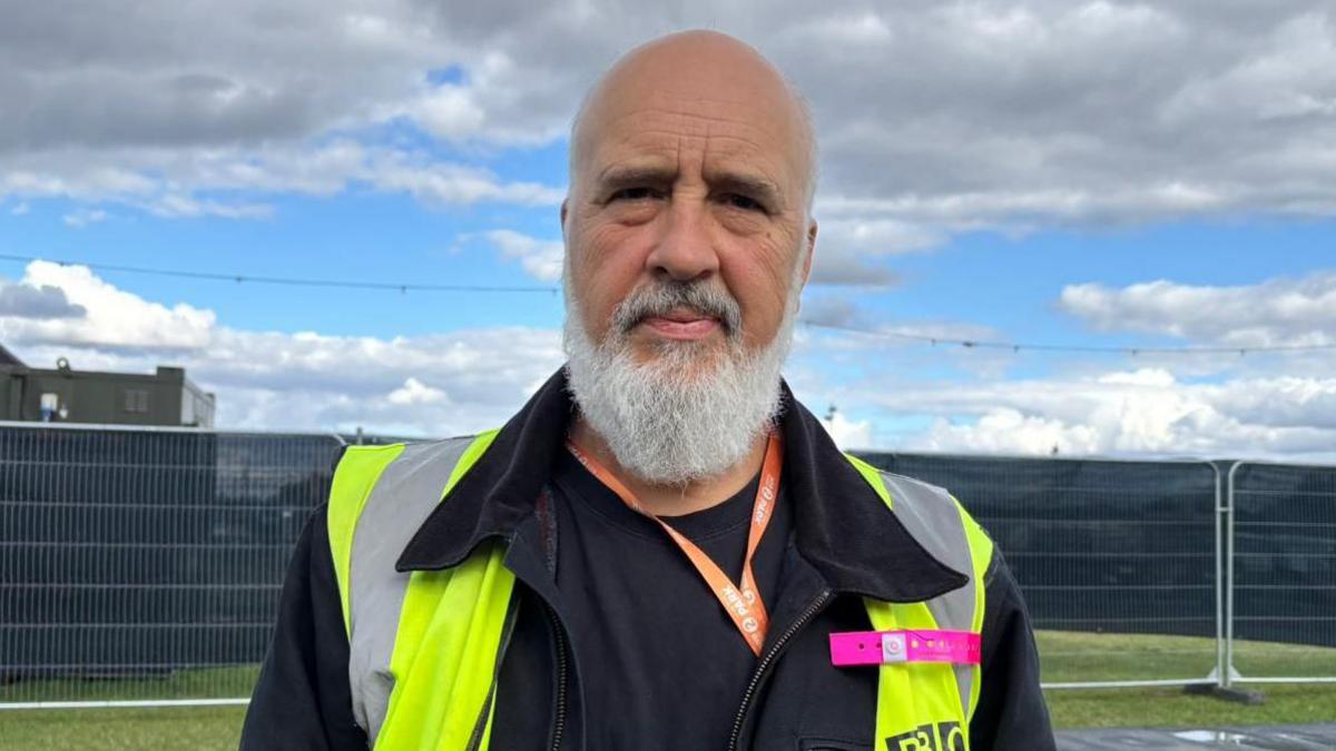 Rhys Hughes looking at the camera while outside at the festival site. He has a bald head and a long grey beard and moustache. He is wearing a hi-vis jacket and a lanyard and there is fencing behind him.