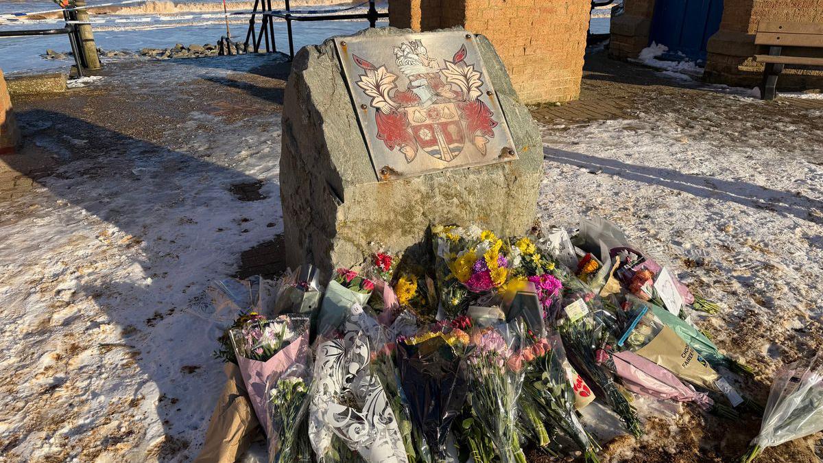 Një pamje nga afër e luleve të vendosura pranë një guri përkujtimor pranë plazhit.