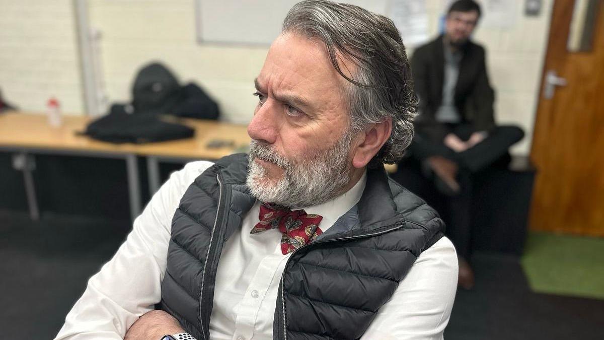 Robert in a rehearsal room, wearing a white shirt, with a bow tie, and a black gilet, with his arms across his chest and a serious look on his face. 