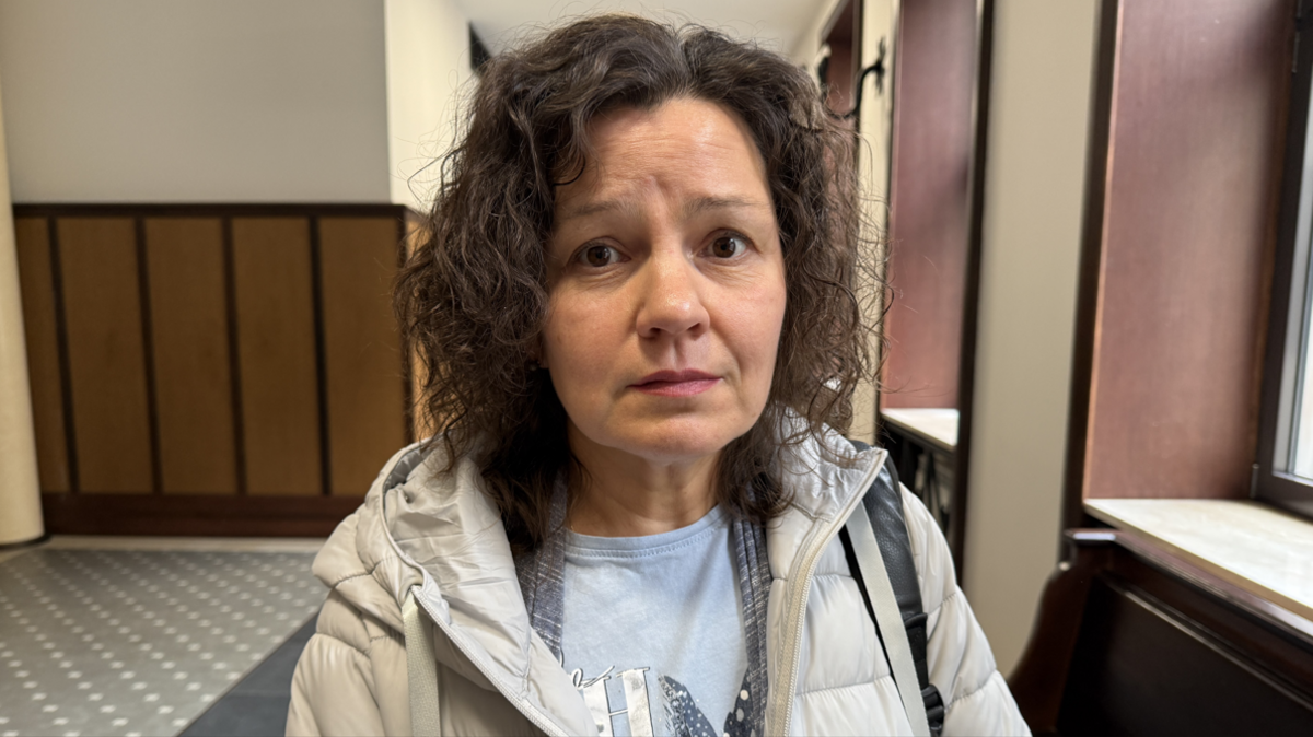 A woman with dark curly hair and a beige coat looks at the camera