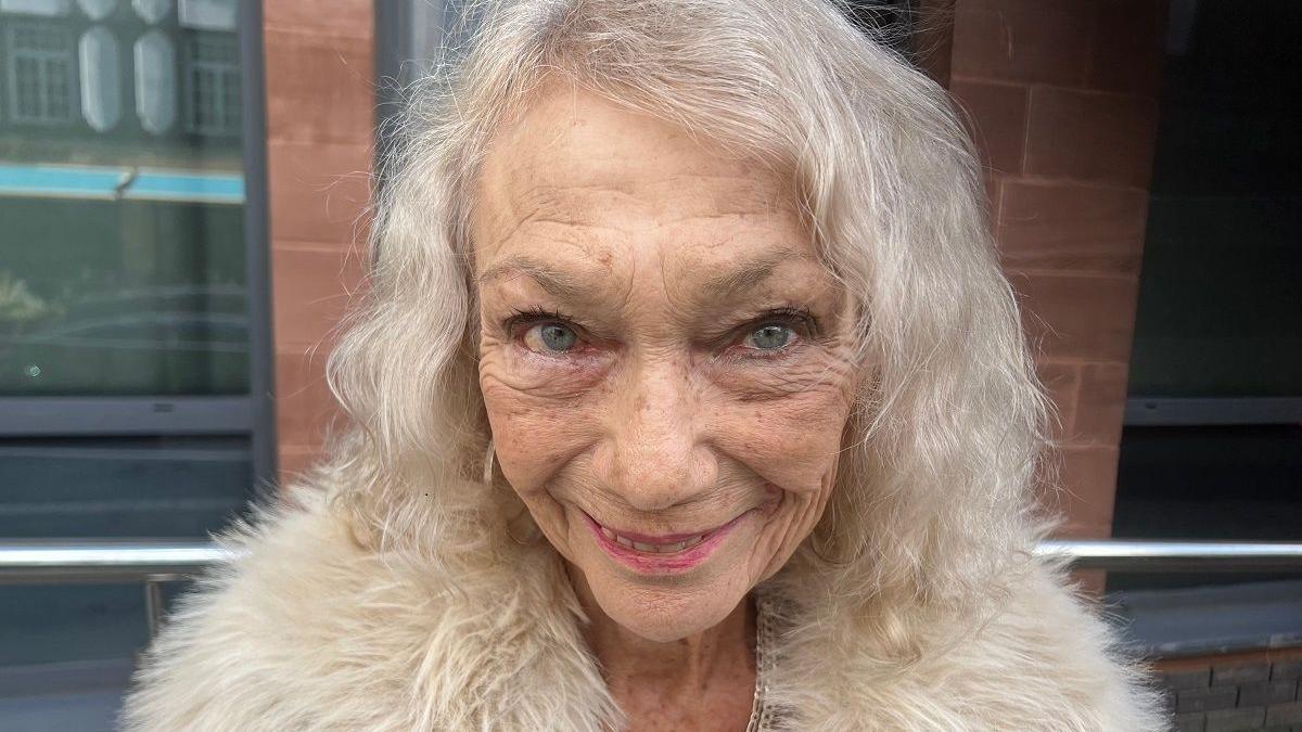 Ellie Mae Constance headshot. she has white hair and is wearing a fur lined cream coat. She smiles at the camera. 