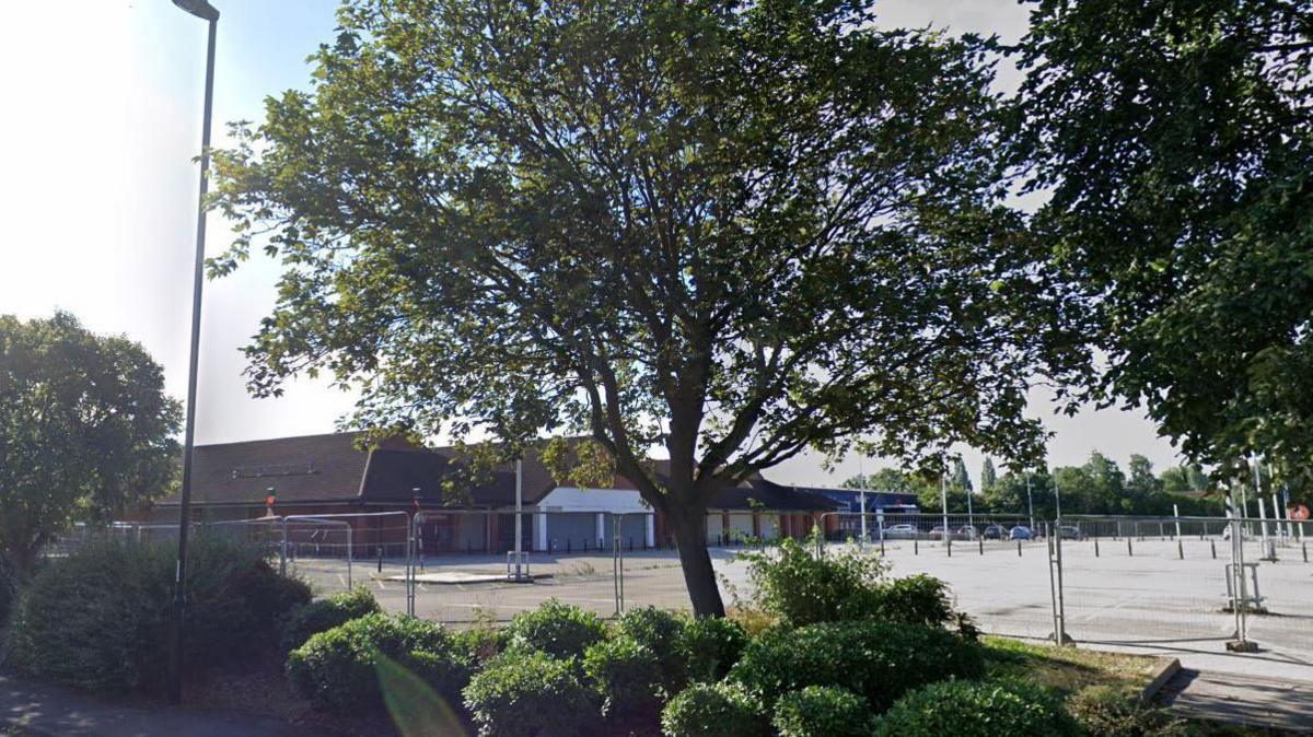 A boarded up supermarket building with barriers around the car park. The site stands empty. There are trees and shrubs planted around the site.