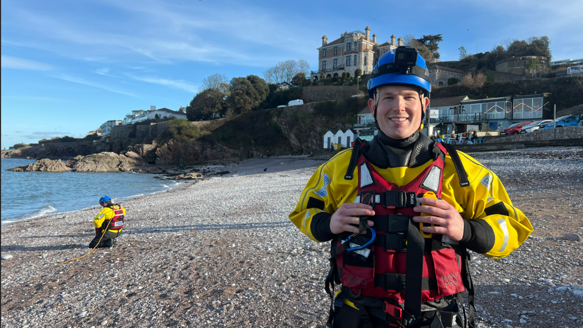 Jack Browse stood at Brixham beach wearing a dry suit. He has a blue hat on, a red vest and yellow jacket. In the background, to the left, is another recruit on the floor. 