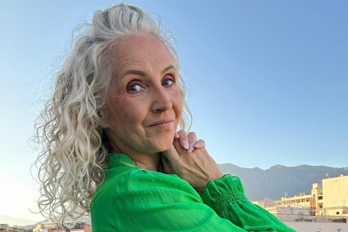 Rhian has curly light blonde/grey hair. In this photographs she is smiling at the camera and wearing an emerald green top. She is in a sunny environment.