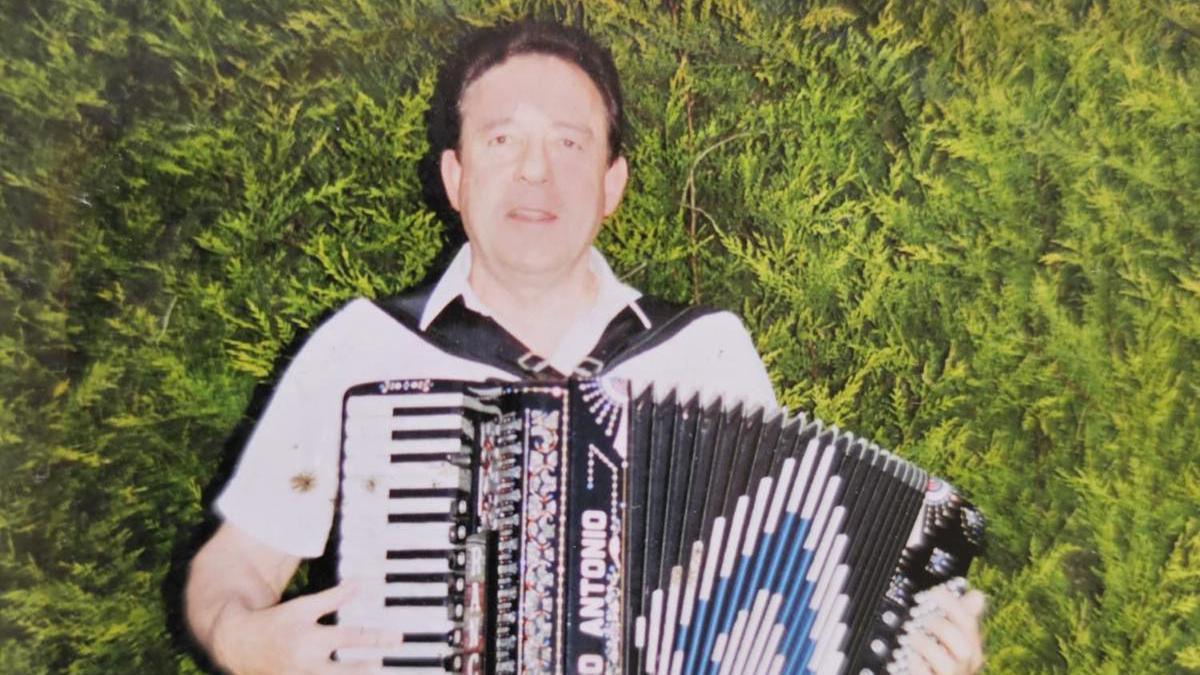 The late Ken Hopkins standing in front of a hedge, playing an accordion. He has short, dark hair and is wearing a white, open-necked shirt.