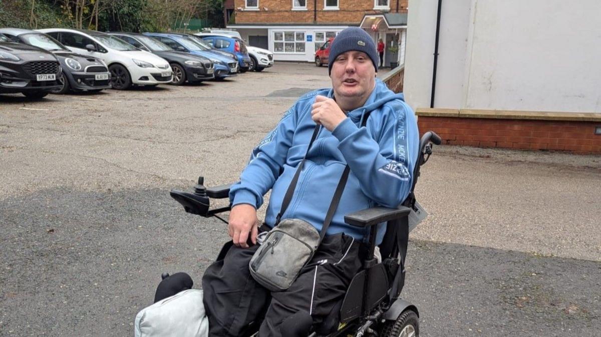 Chris is in a car park in his electric wheelchair. He is wearing a light blue hoody and hat