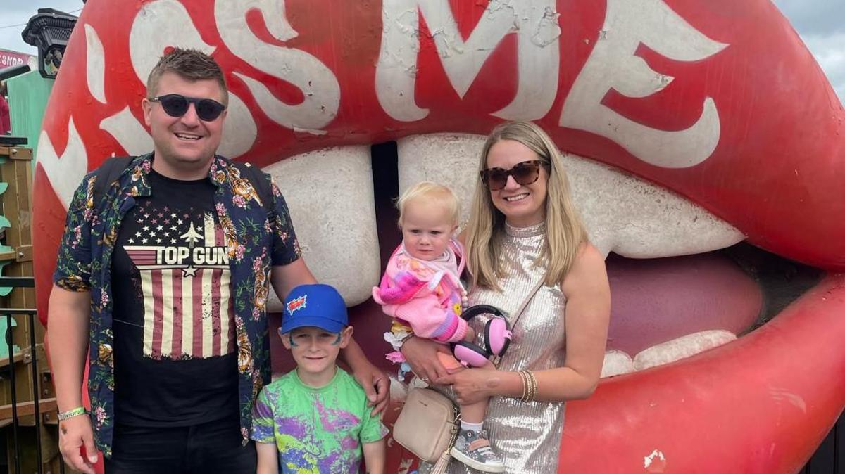 Ben, wearing a Top Gun t shirt and sunglasses, smiling alongside his son Jacob who is wearing a blue cap and a tie dye style green t shirt. Lexi is in her mum's arms, wearing a pink outfit and Melissa is wearing a sparkly silver top. They are in front of a Kiss Me massive pair of lips.