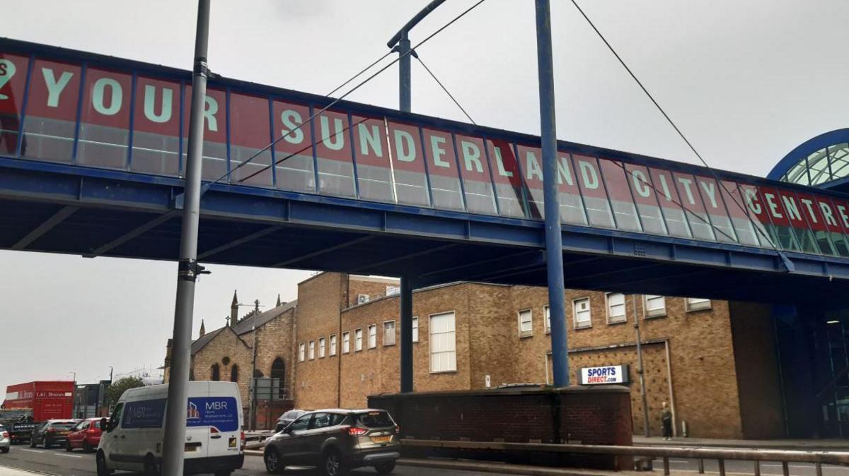 The bridge has a red banner saying Your Sunderland City Centre. It is above a brown brick Sports Direct building and there are cars passing underneath. The bridge itself looks to be enclosed and is made of grey and black metal with large rectangular window panes.