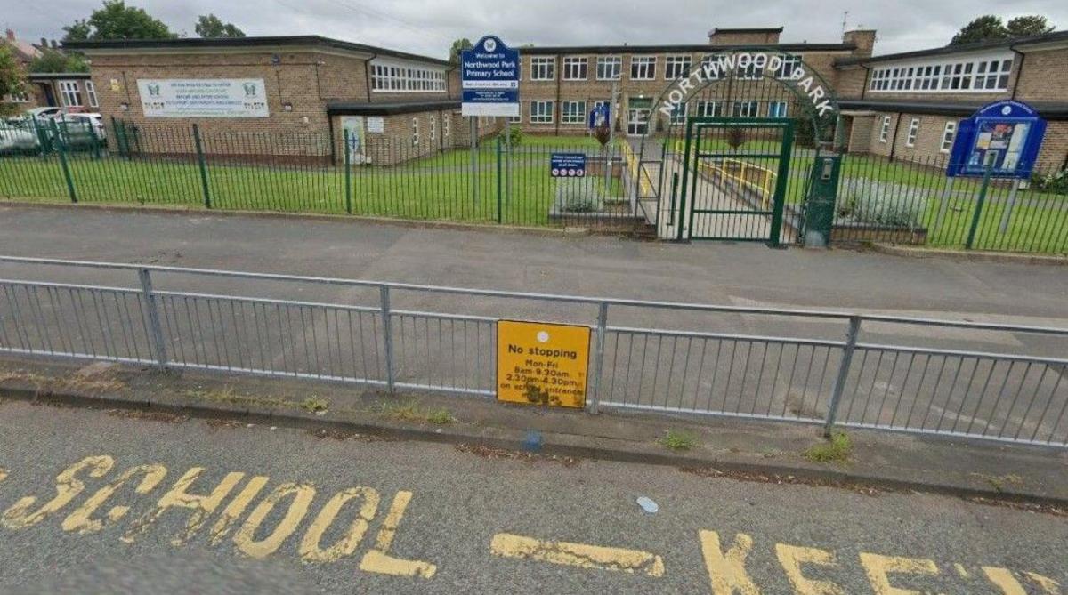 Yellow road markings are on the road in the foreground. The school is behind a fence and the words Northwood Park are above the gate that forms the entrance to the grounds.