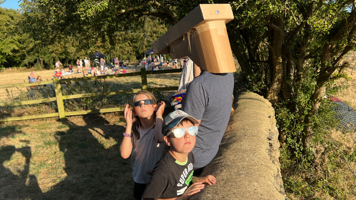 A man is using a cardboard box while sat on a stone wall to look at the eclipse, while two young children are looking up using special glasses.