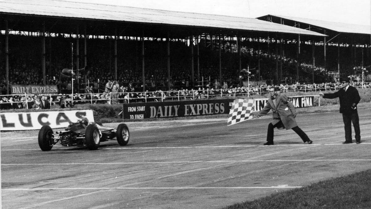 A black and white image of a racing car crossing the finish line at the British GP in 1963. Two men stand on the track close to the winning car, one with a chequered flag, while crowds watch on from the grandstands.