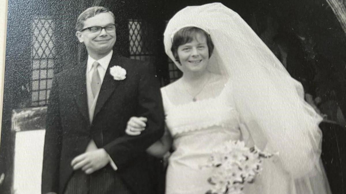 Jan and Peter Cottingham in their wedding outfits in 1966 in a black and white photograph. She has a large veil and is looking at the camera, while her husband is smiling and looking off camera in a dark suit.