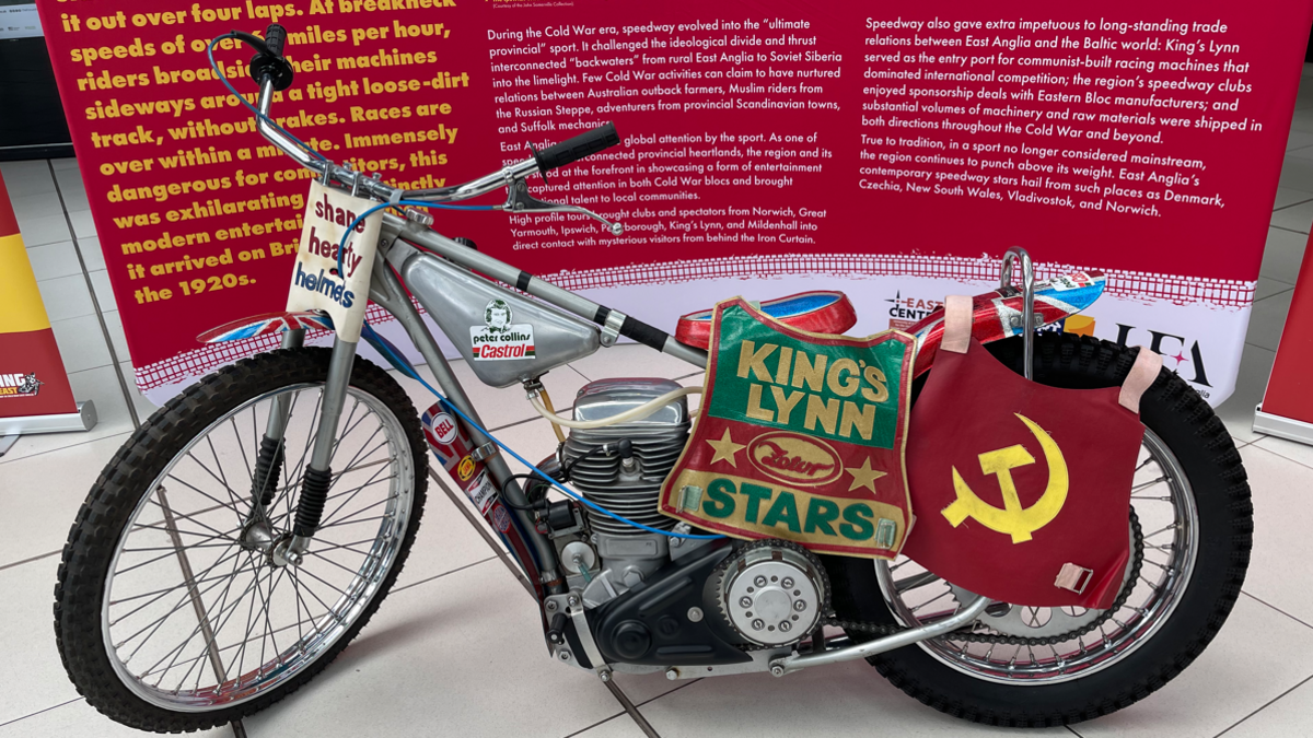 A 1960s speedway bike bedecked with two speedway rider vests - one in red and green with King's Lynn Stars across the chest, and a red one baring the hammer and sickle Communist symbol in yellow 