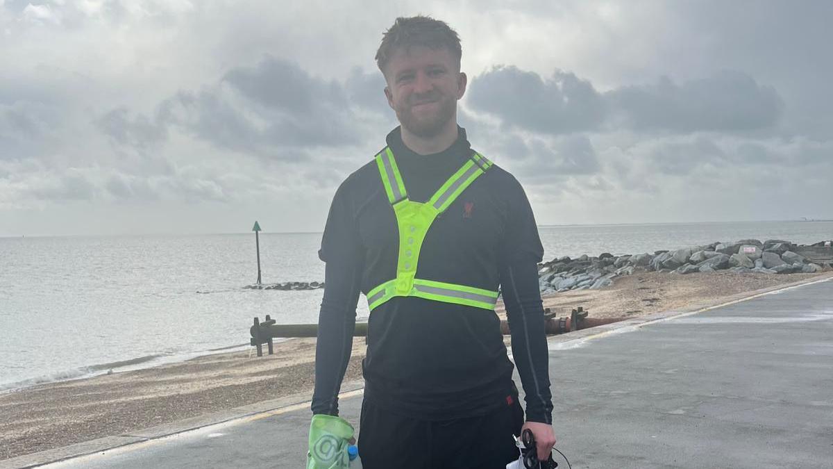 Tommy wearing dark long-sleeved clothing with a reflective harness on. He is standing in front of the sea and a short pebble beach on an overcast day.
