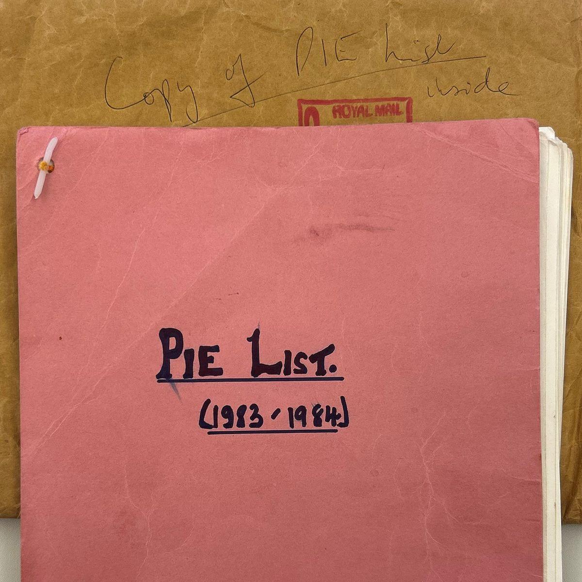 Top down image looking at the pink front cover sheet on the PIE list that Dave Flanagan added when he was in the Met Police. The cover reads in felt tip pen "PIE list - 1983-84". There is a crumpled brown envelope behind, with a red Royal Mail stamp, that reads "Copy of PIE list inside".