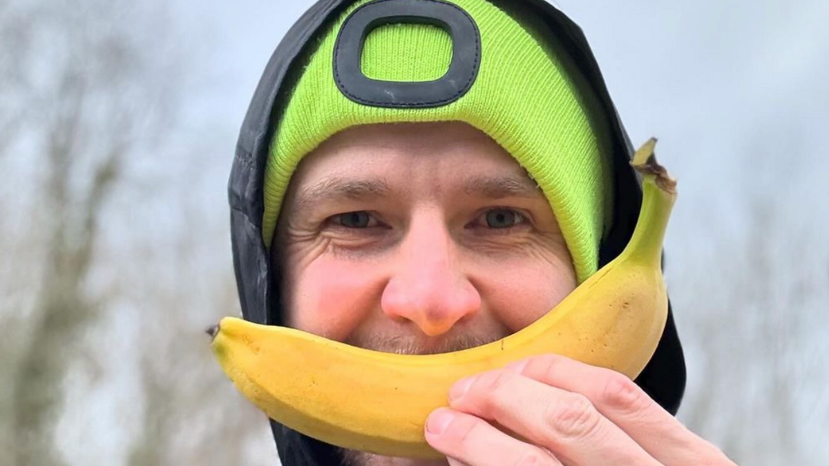 man in beanie hat with hood up, smiling with a banana over his mouth