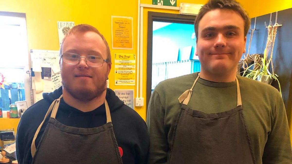 Volunteers Jack and Eden smiling at the camera. They are wearing jumpers and aprons. Jack has short blonde hair and glasses and Eden has short brown hair.