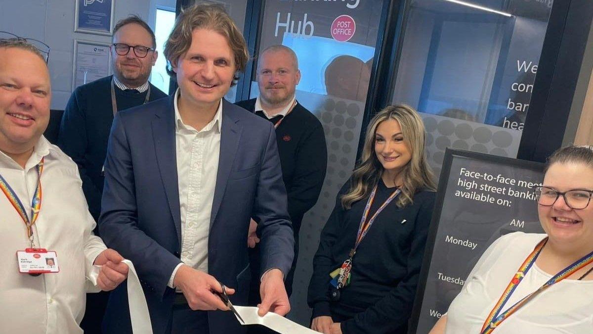 Steff Aquarone, pictured in a banking hub, smiling while holding a pair of scissors and a white ribbon. Five people, two women and three men, stand alongside him smiling.