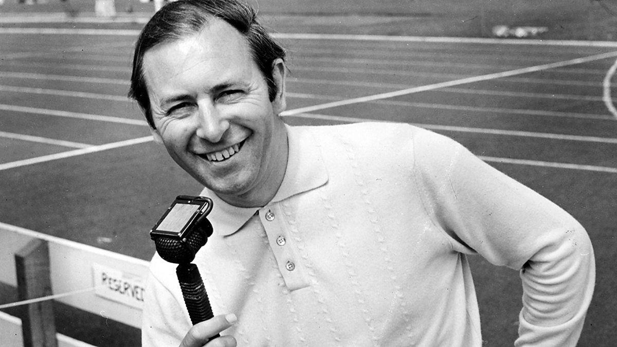 A black and white archived picture of David Coleman as a middle aged man. He is wearing a buttoned-up shirt and has his dark hair parted to the side. He is holding an old microphone in his right hand and standing in front of a football pitch, smiling at the camera.