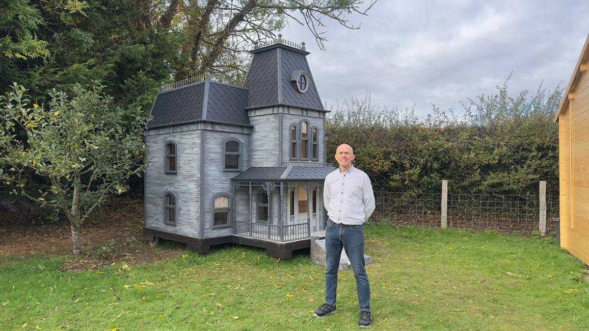 A bald man wearing a grey shirt and blue jeans is standing in a green garden. Behind him, there is a large 12ft grey wooden Halloween house, with sloped roofs and small arched windows