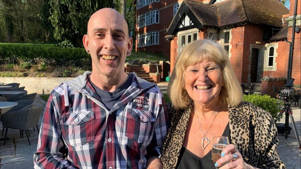 Paula stands with her now adult son in the sunshine holding drinks, both smiling