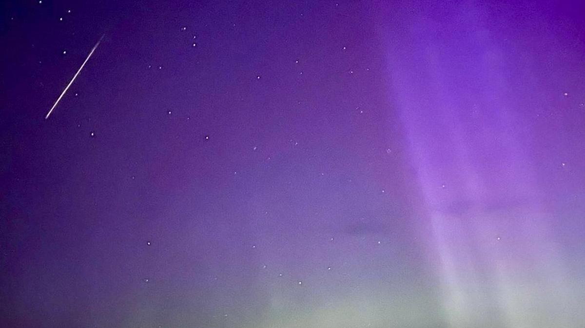 purple night sky with hints of the aurora combined with a few white dots of stars and a streak of white light through the night sky showing a meteor