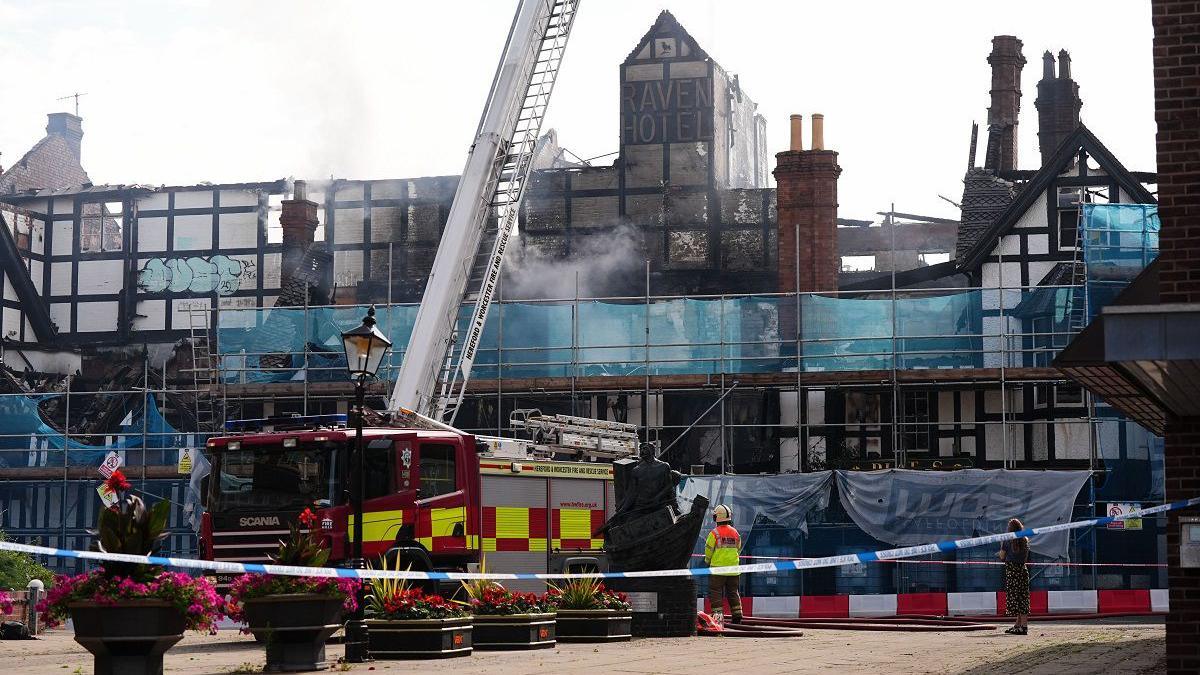 Fire crews are seen working at the Raven Hotel on Monday, the day after the fire started. There is police tape cordoning off the scene and a fire engine has a ladder over the site. The black and white timbered building has scaffolding and parts of it are missing and there is still smoke.
