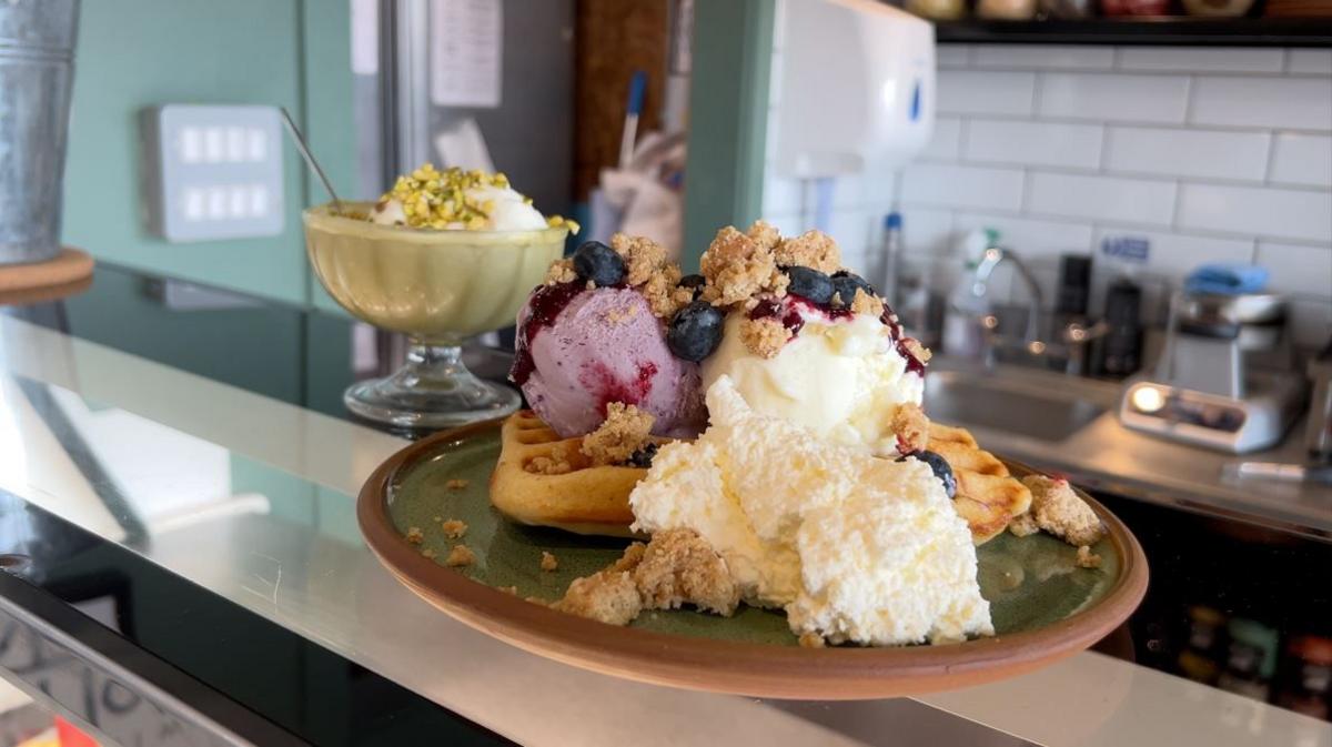 Image of a waffle topped by gelato ice cream blueberries and crumble on a green plate on a cafe counter top. 