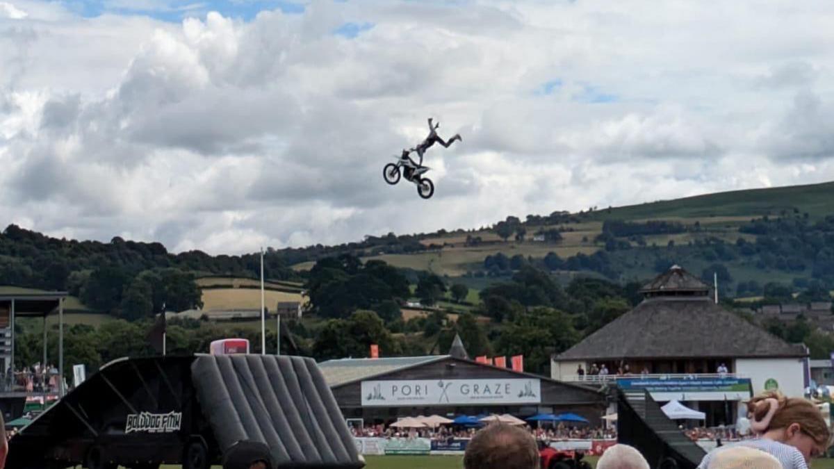 A man doing a dramatic stunt in the air above a field in the festival. The driver is holding onto his bike whilst his legs are in the air. Crowds can be seen watching the stunt. 