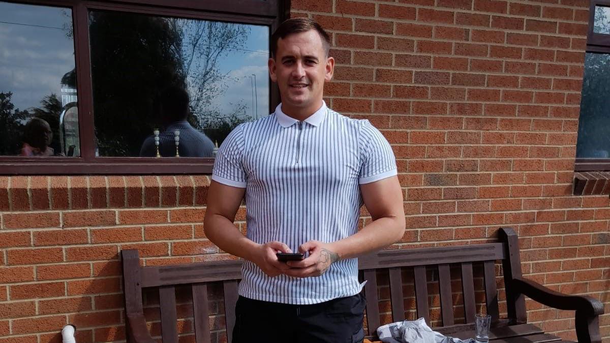 A man in a blue and white striped zip polo holds a phone and stand in front of a brown wooden bench smiling at the camera.