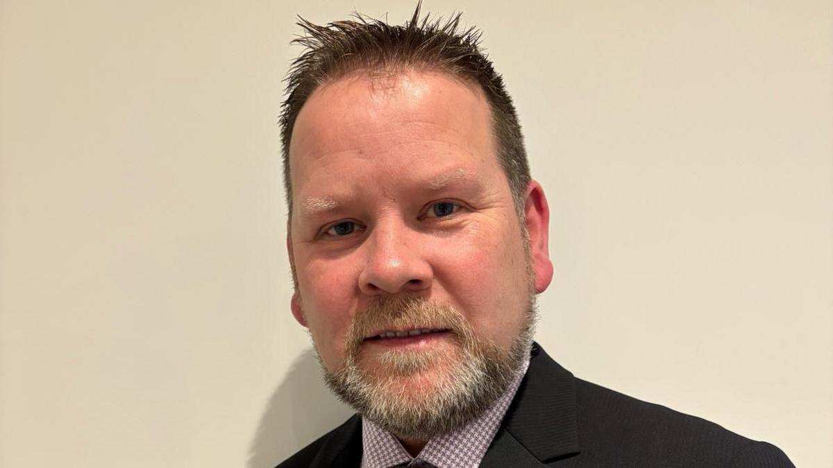 A man with brown hair and a grey beard looking into the camera he is wearing a purple shirt and a black blazer suit. he has blue eyes . 