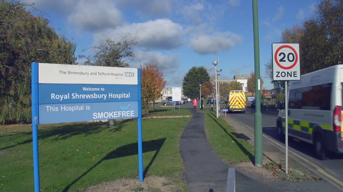 The entrance to a hospital with an ambulance passing a blue and white sign on a grass verge with the words Welcome to Royal Shrewsbury Hospital written on it