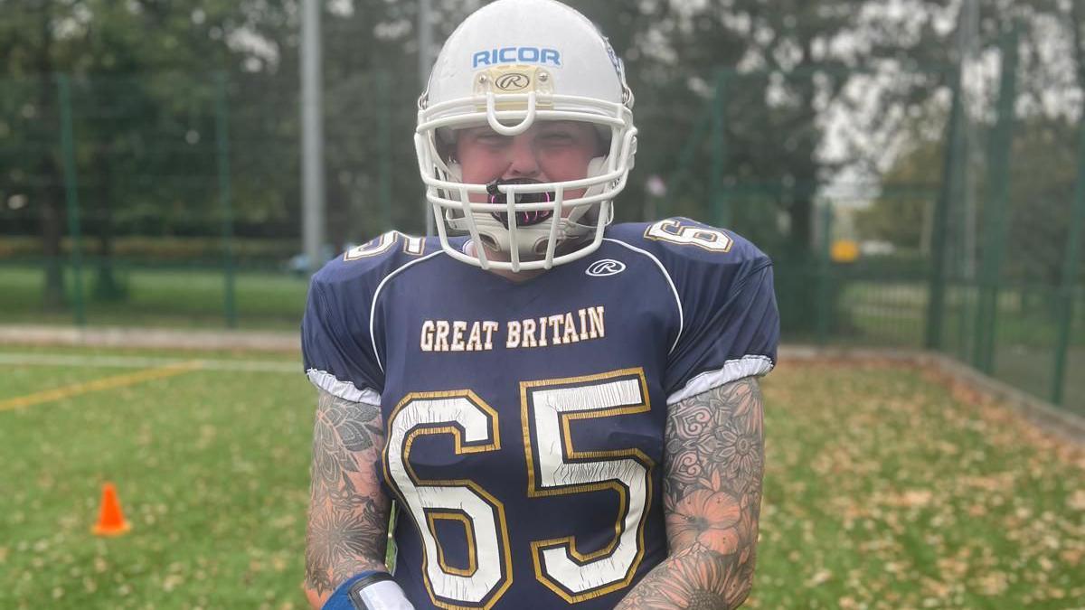 Jenna Abbott smiling and wearing a navy American football jersey with white letters that say 'Great Britain' on the front and the number 65 which has a yellow outline. She is wearing a white helmet.