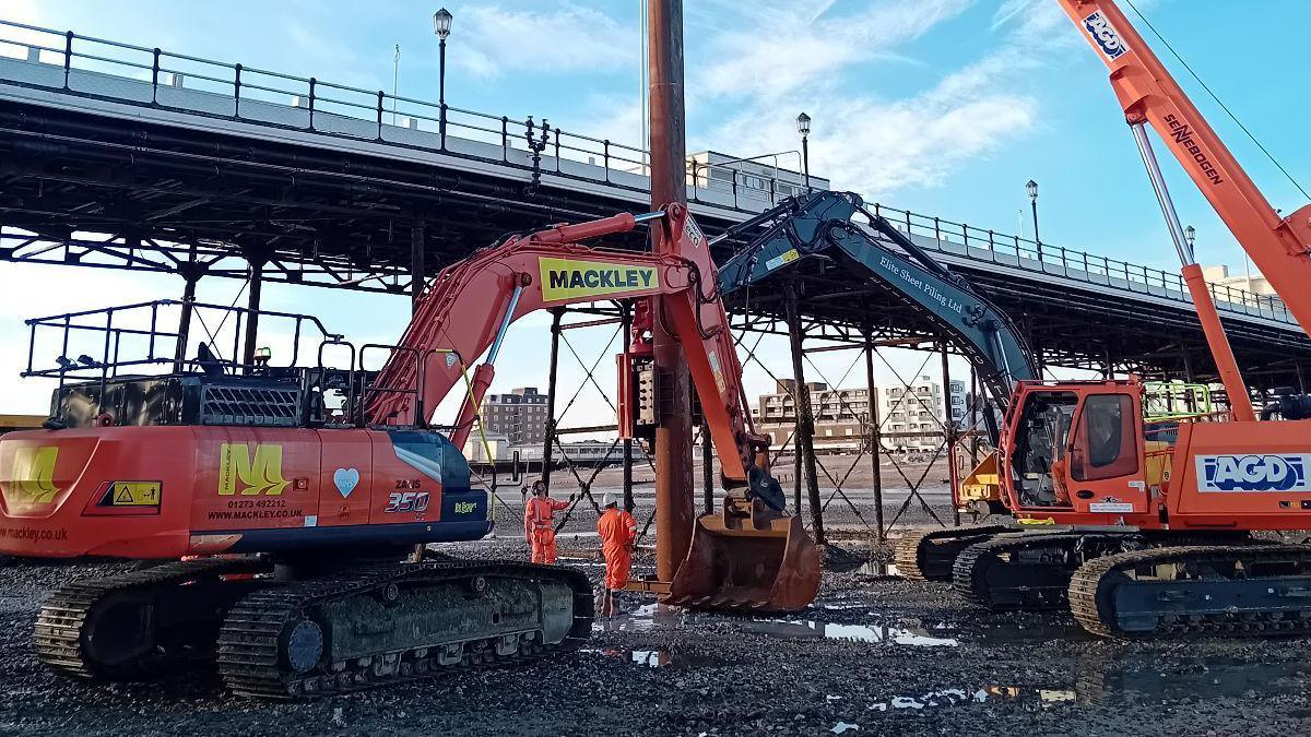 Two large diggers on the pebbles on the beach in Worthing. Two workers in orange high-vis outfits are stood next to them, under the pier. 