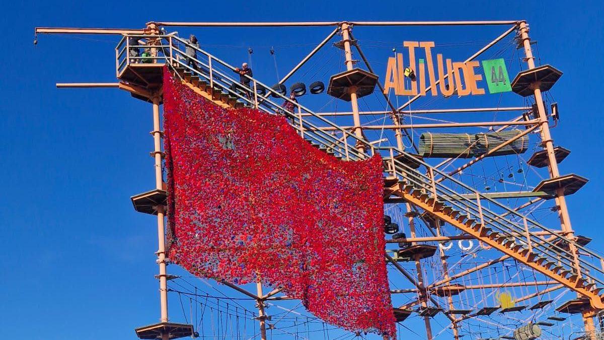 A tall attraction ride with several levels which says 'Altitude 44' in orange and green text at the side. A large rectangular flurry of red poppies are draped over the side, with a blue sky in the background.