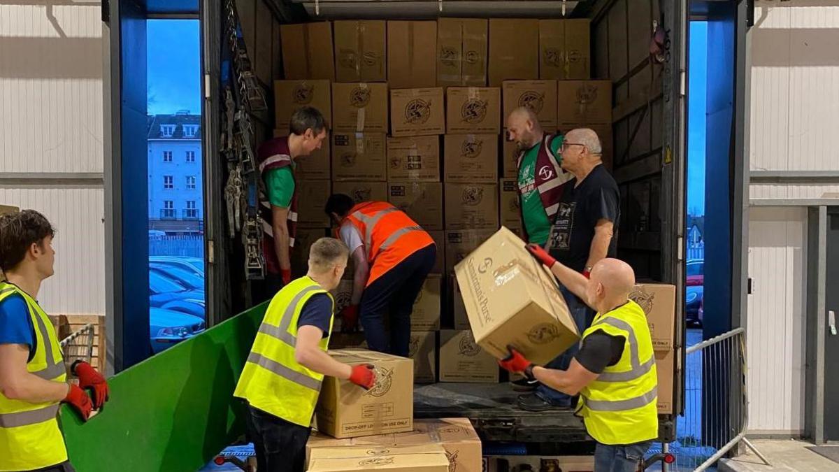 Volunteers loading brown cardboard boxes into a lorry. They are all wearing Hi-Viz jackets, three in yellow, one in orange and two in burgundy. 